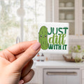 A hand holding a sticker with a cartoon image of a pickle and the text "Just dill with it" in a kitchen setting.
