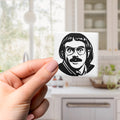A hand holding a small square magnet with a black and white illustration of a man's face on it.