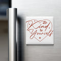 A white heart-shaped magnet with the text "Be Kind to Yourself" written on it, placed on the surface of a stainless steel refrigerator.