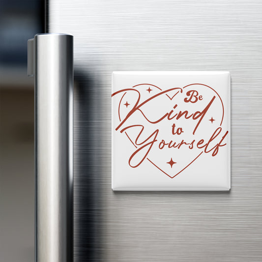 A white heart-shaped magnet with the text "Be Kind to Yourself" written on it, placed on the surface of a stainless steel refrigerator.