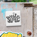 A white square magnet with the text "sweater weather" written on it, attached to a weathered concrete surface.